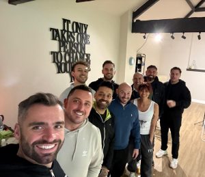 A group of smiling people in a room with a wall sign reading, It only takes one person to change your life