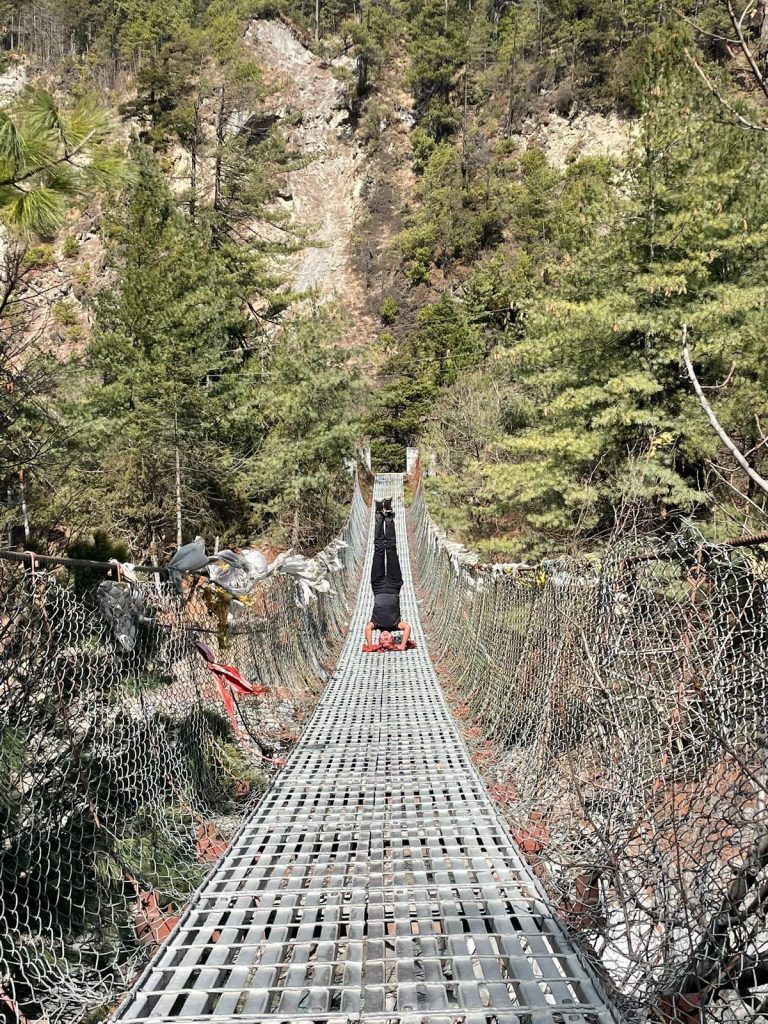 A person performing a headstand on a long suspension bridge surrounded by a forested mountain slope