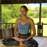 Woman meditating in a cross-legged pose on a woven mat in an open-air wooden structure
