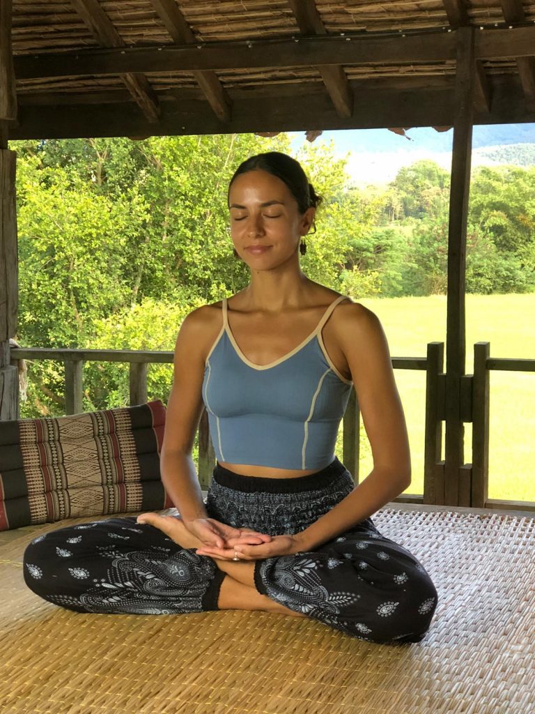 Woman meditating in a cross-legged pose on a woven mat in an open-air wooden structure