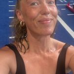 A smiling, sweaty woman in a black tank top sitting on a blue turf track in a gym